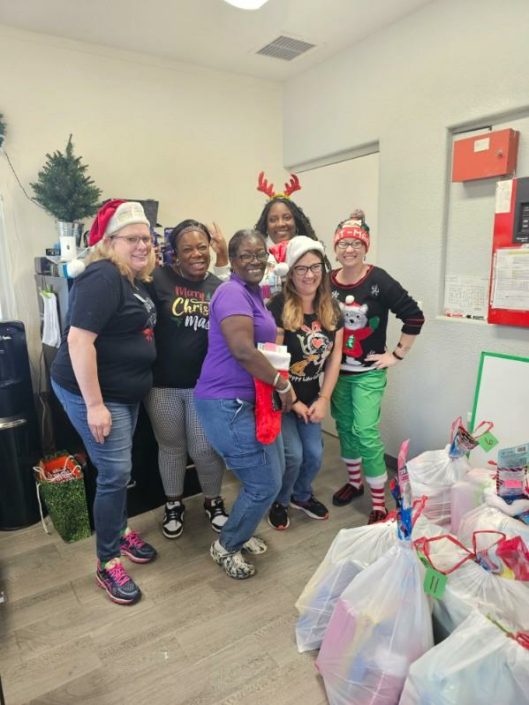 Six women wearing holiday sweaters and Santa hats, posing with Christmas gifts.